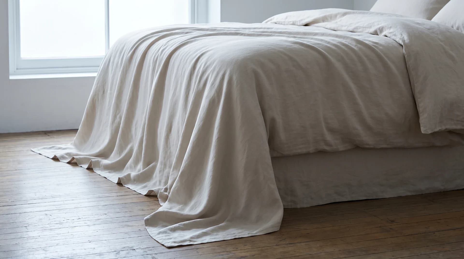 A bed dressed in smooth pale bedding, one sheet falling loose and pooling on the wooden floor, in cool daylight from a window.