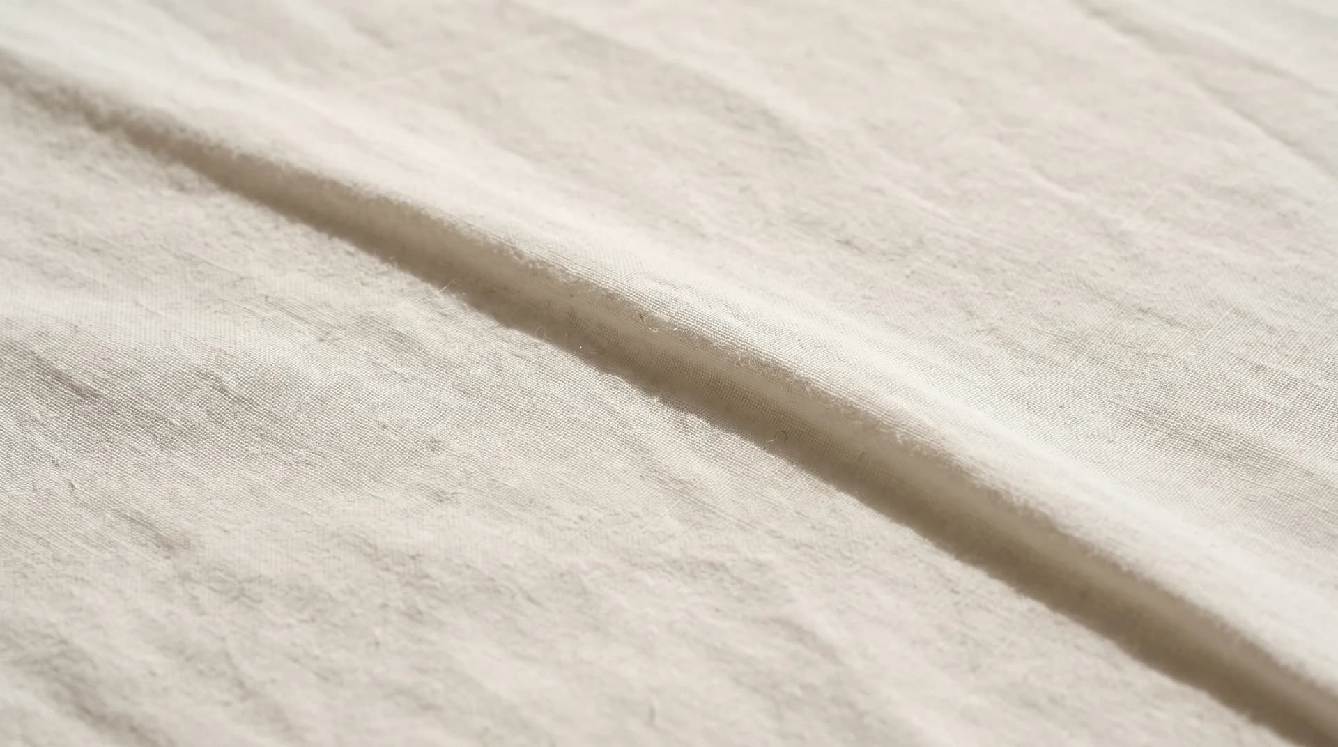 Close view of pale cotton sheeting folded across itself, its surface catching soft directional light.