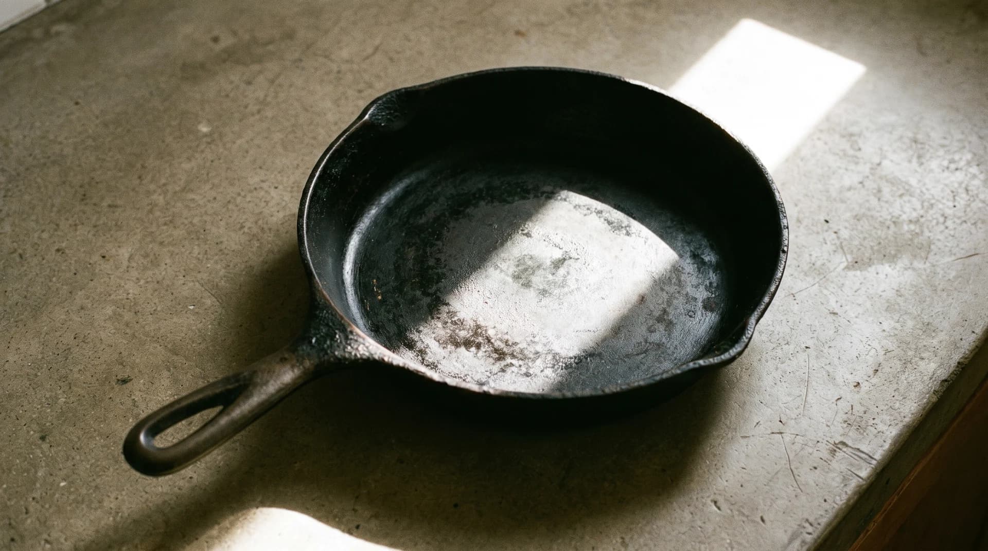 A well-used black cast iron skillet on a concrete counter, a hard shaft of daylight falling across its dark, unevenly patinated cooking surface.