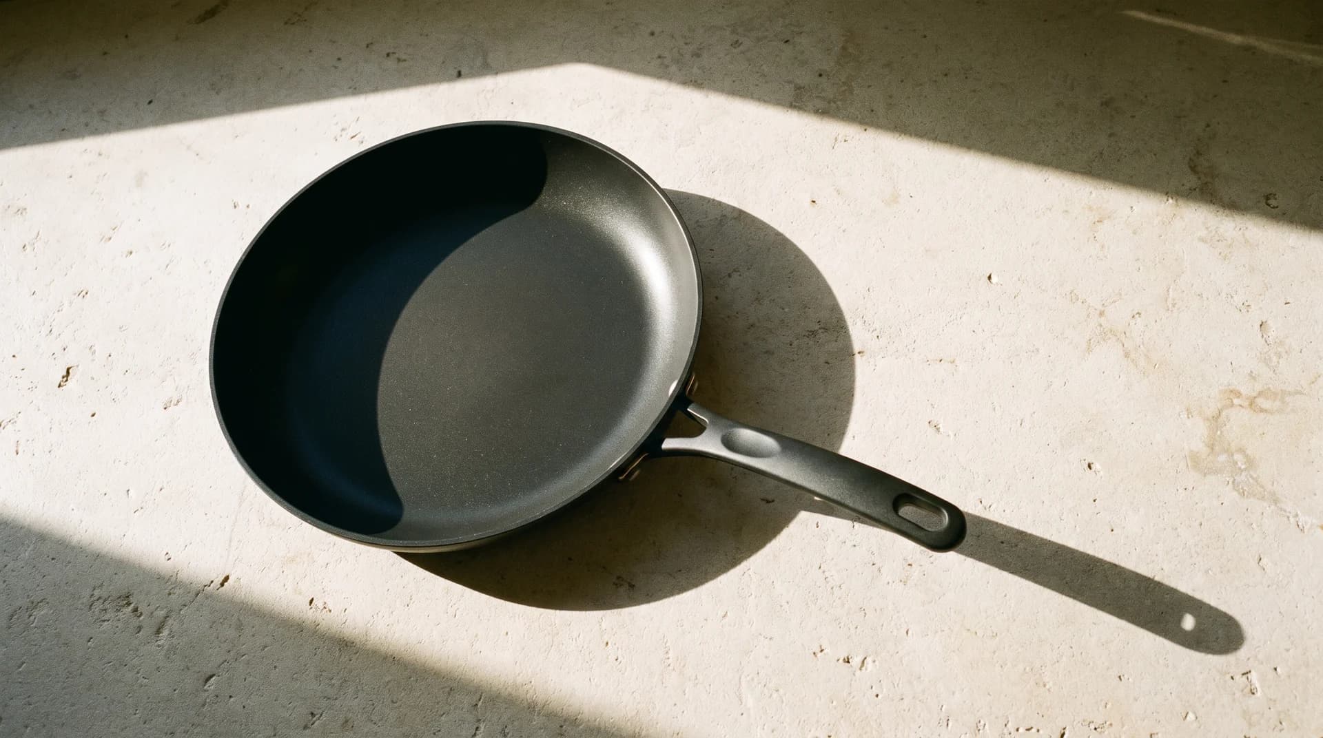 A dark nonstick frying pan on a pale stone counter, lit from one side so it casts a single long shadow across the stone.